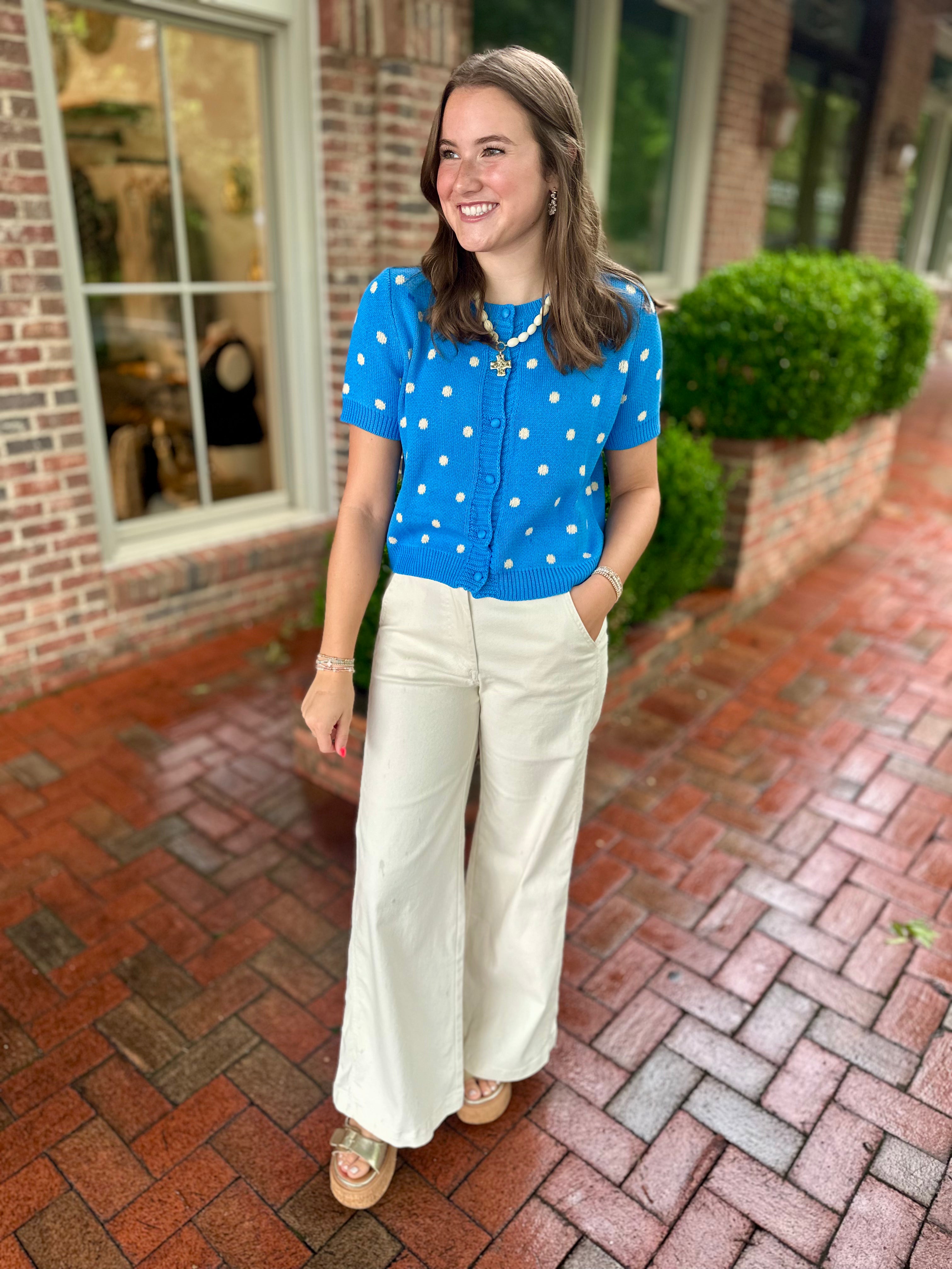 Model wearing a bright blue short-sleeve polka dot cardigan paired with cream wide leg pants from Pink Chair Boutique.